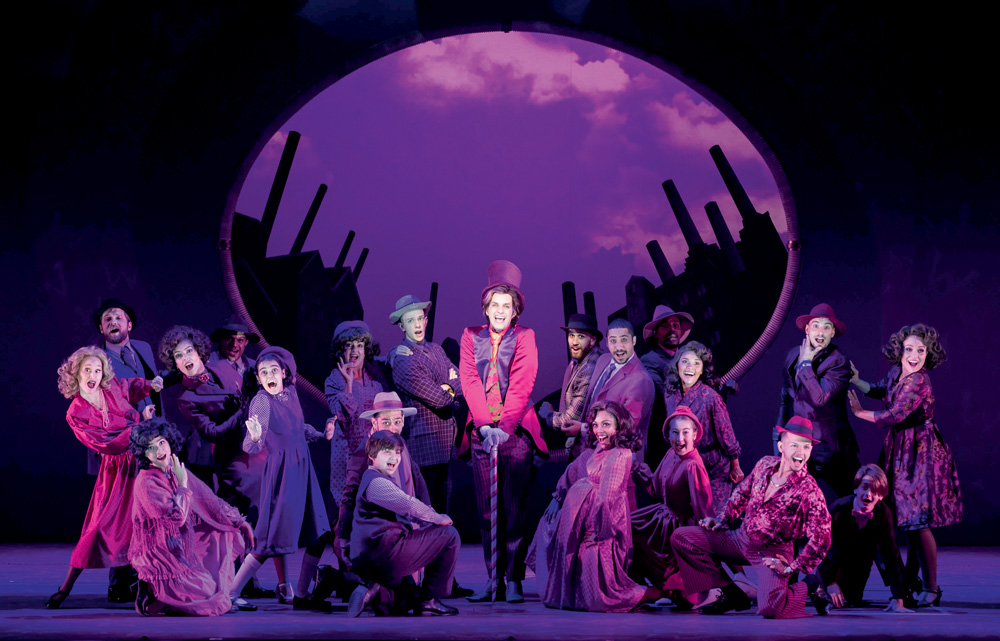 Mais de dez atores, com roupas roxas, crianças e adultos, estão em um palco de teatro, roxo e com iluminação roxa. O homem no centro está vestido - e interpretando - o Willy Wonka