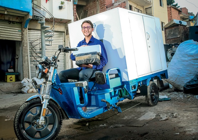 Um jovem está em uma moto que carrega encomendas (com um grande suporte atrás), em uma favela. Ele sorri para a foto