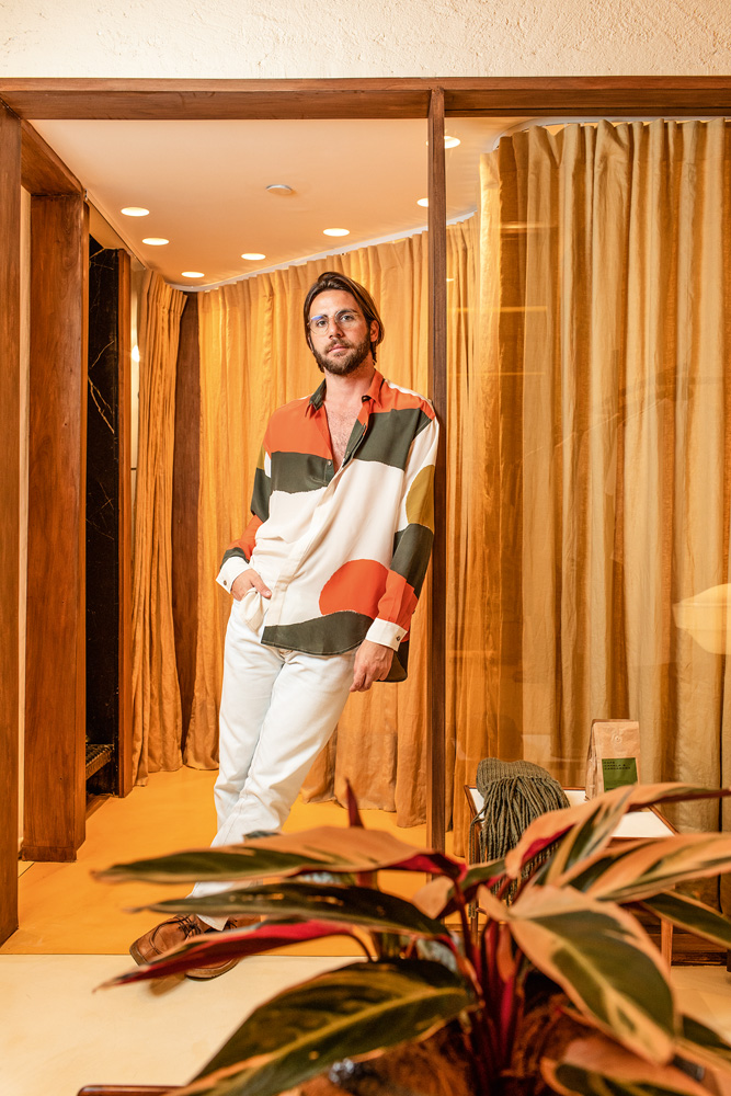 HANDRED18.jpg Um homem vestido de camisa e calça branca, com detalhes em verde e laranja, posa no provador de uma grife no shopping