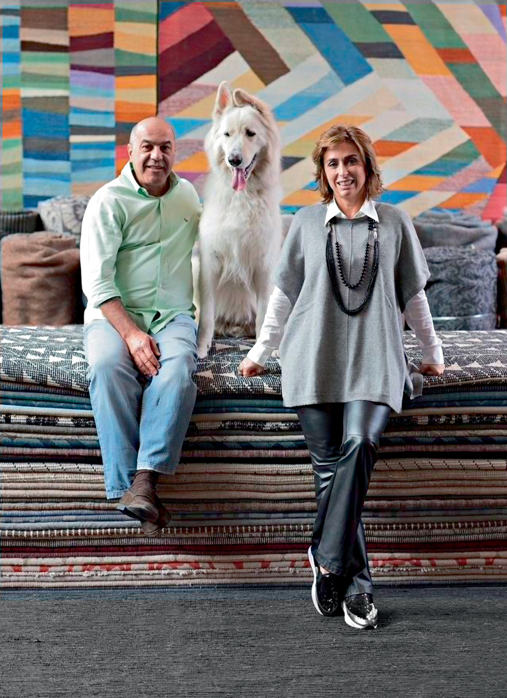 Kamyar-Abrarpour-Francesca-Alzati-fundadores-by-Kamy-tapeçaria-obras-de-arte Kamyar Abrarpour e Francesca Alzati posando para foto junto com um cachorro e tapetes da loja, empilhados. kamyar está junto com o cachorro sentado nos tapetes e francesca está apoiado nos tapetes, ao lado de kamyar e o cão