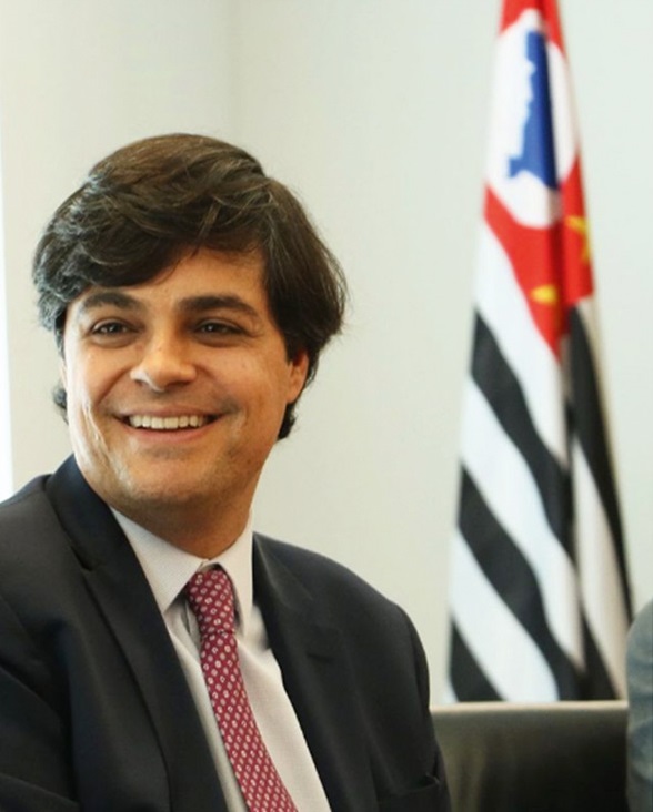 Um homem de terno e gravata sorri e olha para o lado. No fundo da foto, uma bandeira do estado de São Paulo