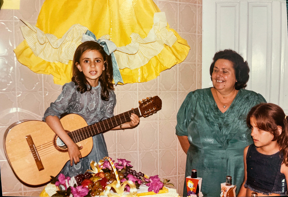 A imagem mostra Ludmilla com 7 anos, em seu aniversário, tocando violão enquanto é observada pela avó.