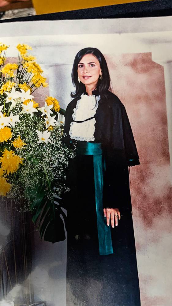 A imagem mostra Ludhmilla, com roupa de formatura, ao lado de flores.
