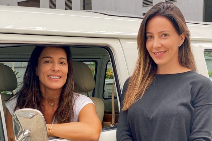 A imagem mostra Mariana, dentro de um kombi no banco do motorista com o cotovelo para fora do veículo. Do seu lado, Carolina, com um mão segurando o pulso da outra. As duas estão sorrindo para a câmera.