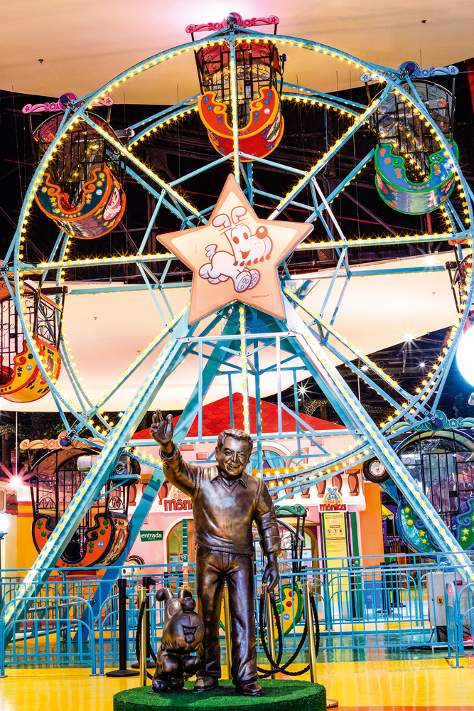 Uma foto de uma roda-gigante estampada com vários personagens da Turma da Mônica. Na frente do brinquedo, uma estátua em bronze de Mauricio de Sousa