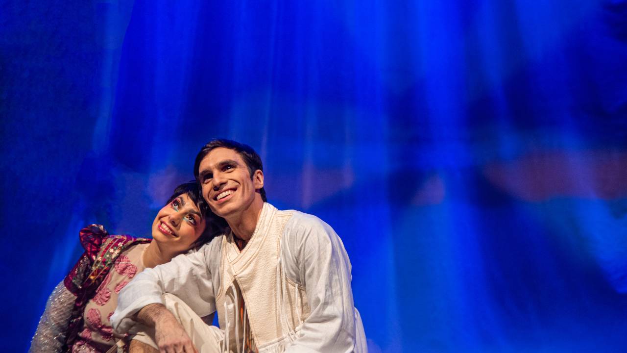 Em um palco de teatro com luzes azuis bem fortes, estão um jovem e uma jovem sentados juntos olhando para o que seria o céu