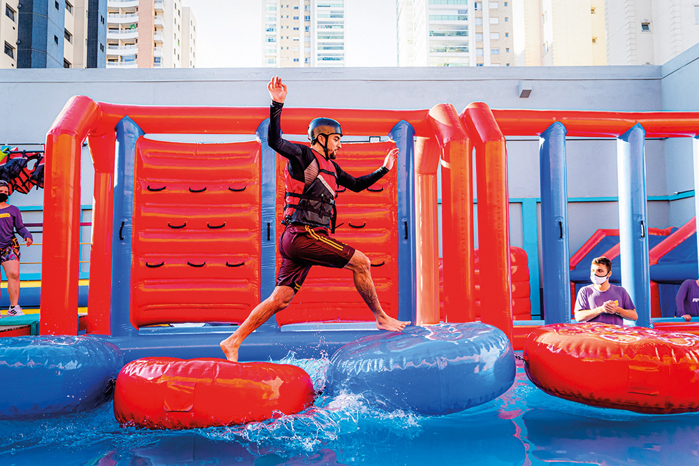 Em um parque de brinquedos infláveis, um jovem está pulando em um deles. Ele pula entre duas boias que estão em uma piscina. Boias azuis e vermelhas