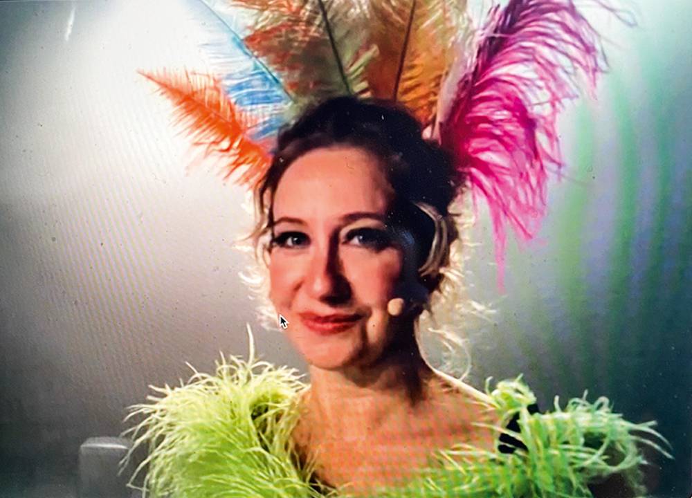 Mulher loira olha para a câmera sorrindo. Ela está maquiada e de microfone. Está também com penas na cabeça e roupa verde