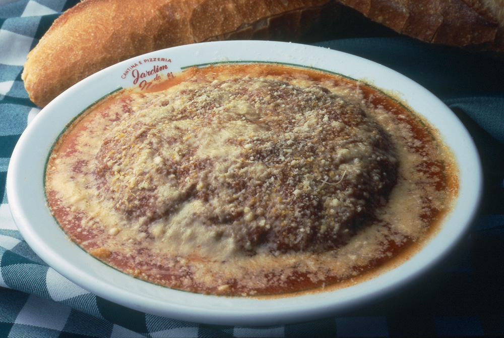 Bolo de carne achatado e frito e recheado de muçarela no molho de tomate conhecido como polpettone alla parmigiana sobre um prato raso de cor branca