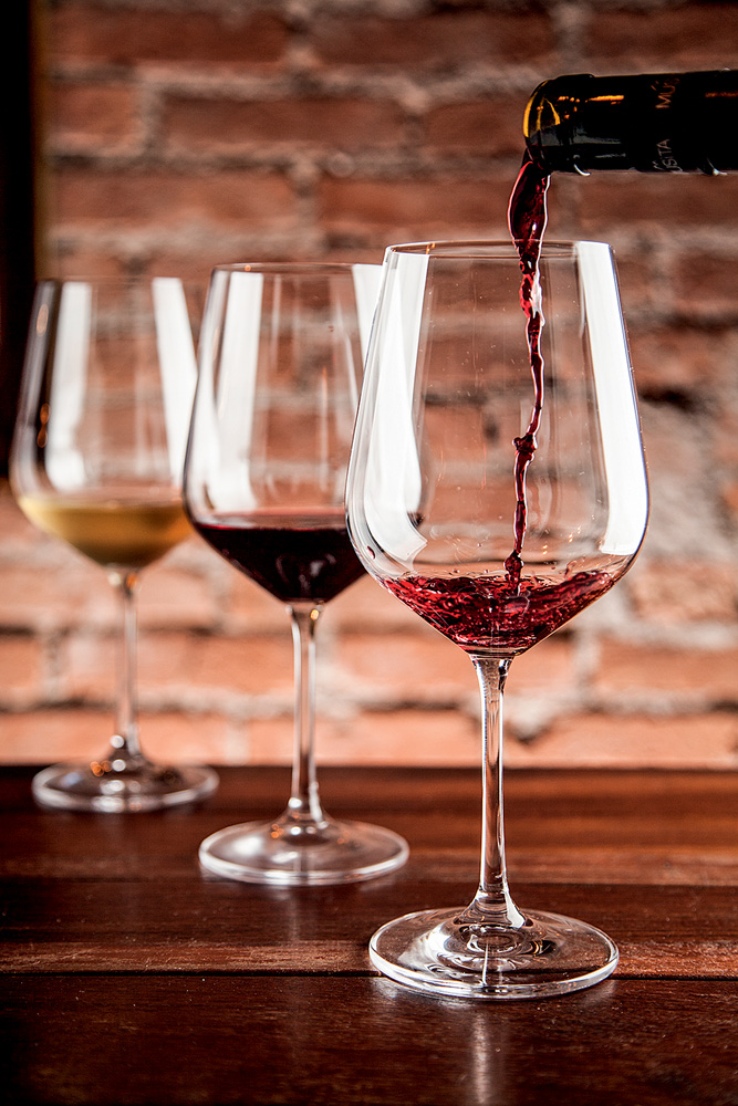 Três taças de vinho alinhadas em diagonal sobre mesa de madeira. Ao fundo com vinho branco, no meio com tinto e na ponta com tinto sendo servido.