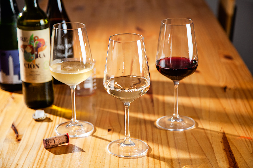 À esquerda, há três garrafas de vinho. Ao lado, três taças de vinhos, dois brancos e um tinto.
