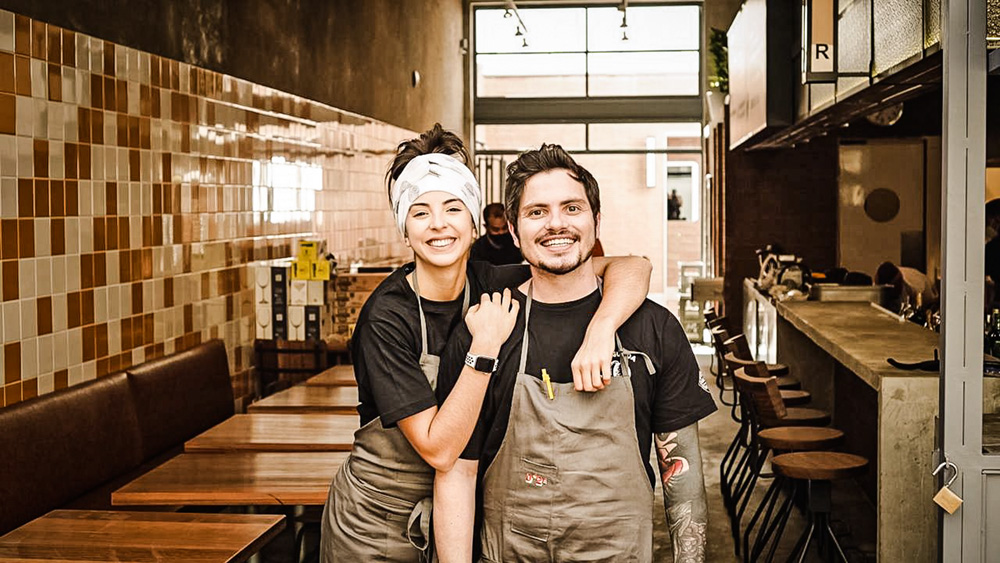 Chefs Júlia Tricate e Gabriel Coelho posam lado a lado em frente ao seu mais novo restaurante, o De*Segunda