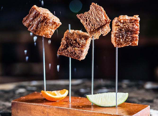 Green Kitchen Porção de quatro unidades de torresmo vegano posicionados sobre pequenos espetos e servidos junto de fatias de limão