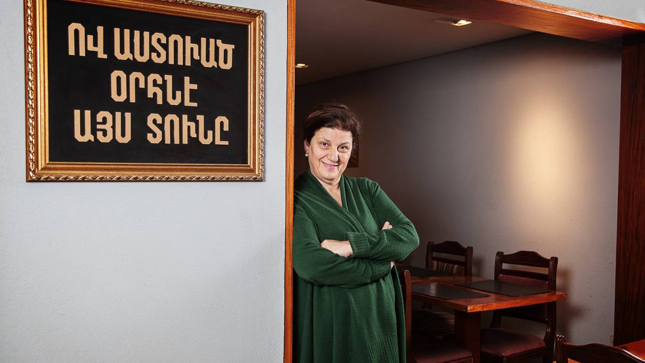 Yeran Garabedian Habib de camisa verde escura encostada na parede de seu restaurante com os braços cruzados.
