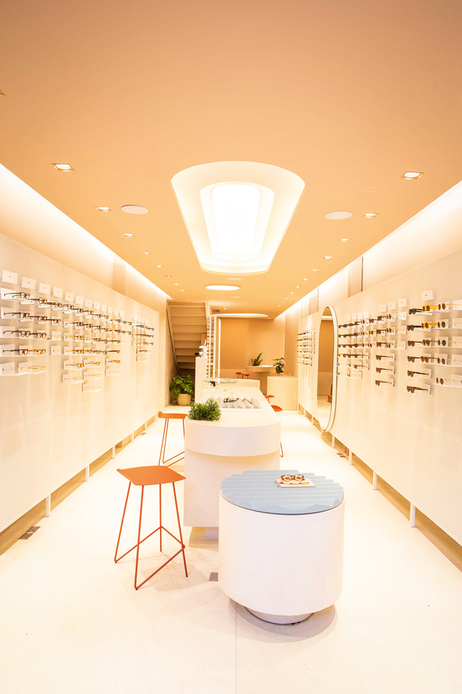 Uma loja de óculos. Paredes brancas com os modelos pendurados, alguns banquinhos e uma mesa no centro