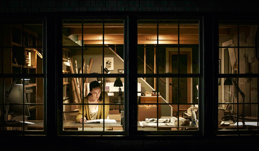A imagem mostra uma mulher, durante a noite, lendo um livro com apenas um abajur e poucas luzes acesas.