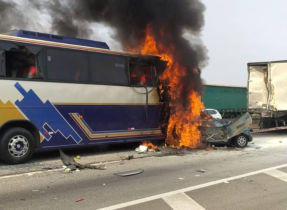 A imagem mostra um ônibus colidido com um carro. Os veículos estão em chamas