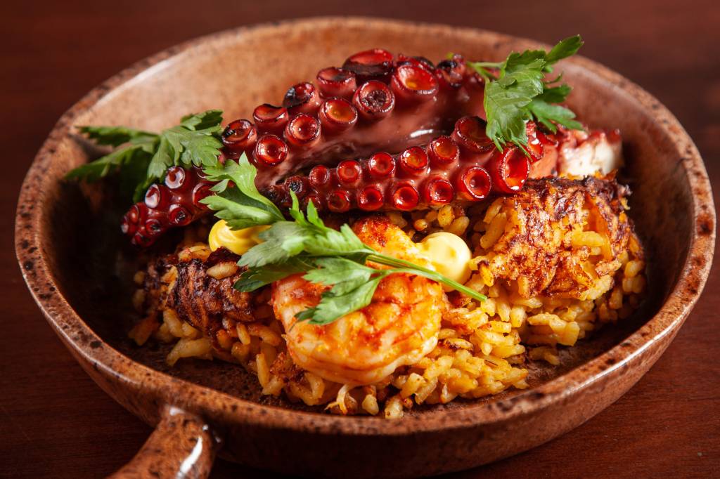 Me Vá Em uma cumbuca funda e de cor marrom, é servido o prato junto de arroz, camarões e dois tentáculos de polvo