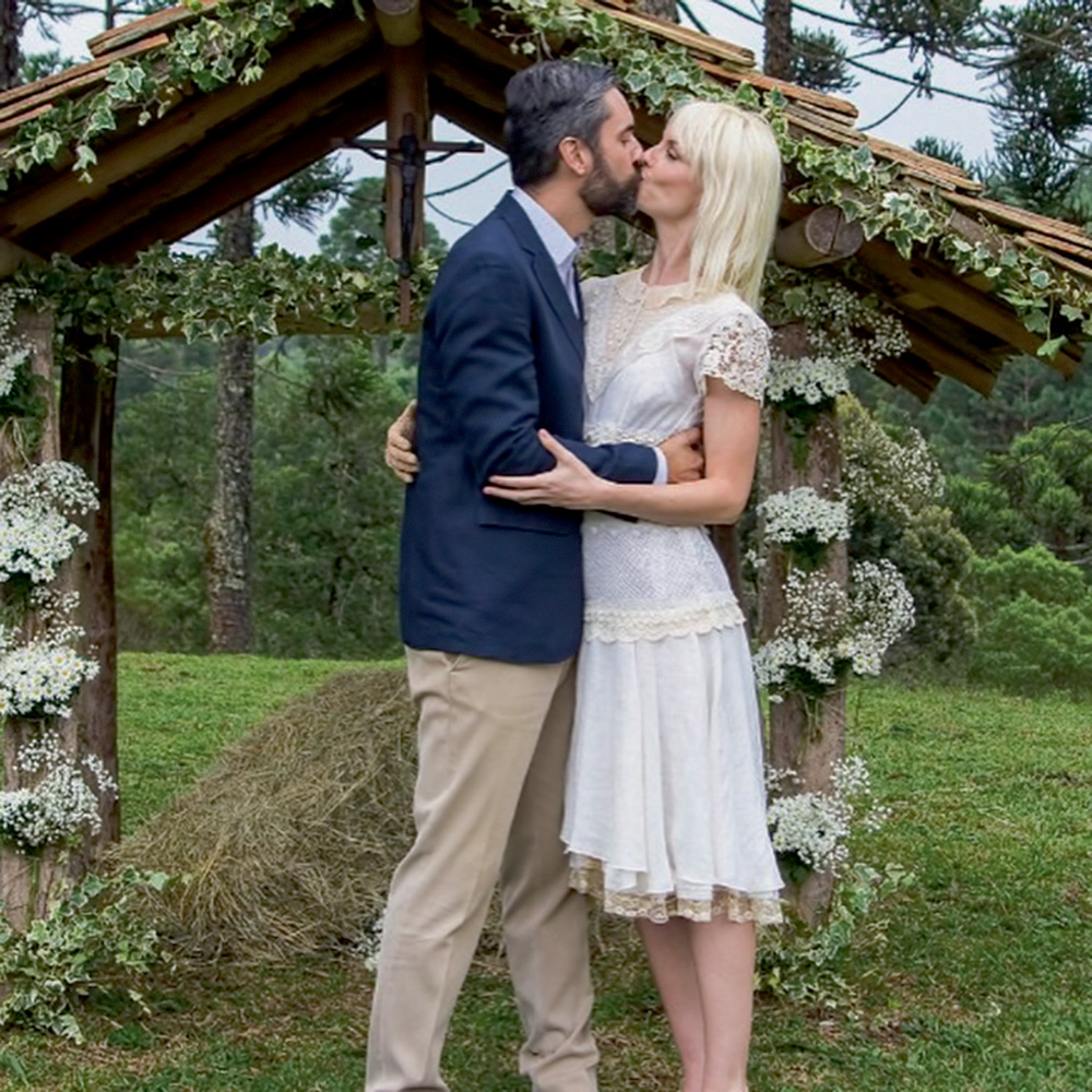 augusto e ana se beijando, ele com um terno, ela de vestido branco, em seu casamento, em um local aberto com gramado e diversas árvores. atrás deles há uma estrutura estilo portal de madeira ornada com flores