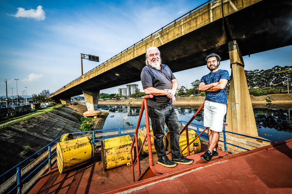 Douglas, 57, e Victor, 37, posando para a foto sobre barco Almirante do Lago. Douglas está à esquerda encostado em um corrimão enquanto sorri e Victor está de braços cruzados e apoiando em outro corrimão, sorrindo. Atrás deles, o rio tietê e um dos viadutos do cebolão