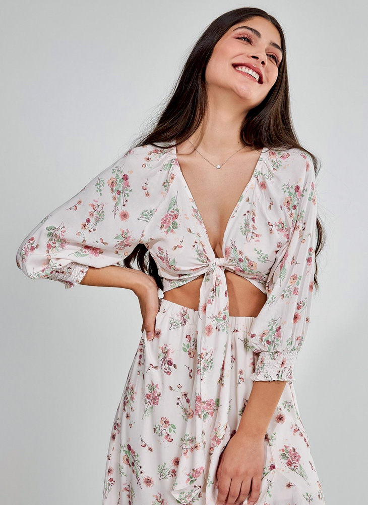 Uma menina, em um fundo branco, sorri com a mão na costela. Ela veste um conjunto com estampa igual, de florzinhas delicadas rosa e verde. O conjunto é uma camisa de amarração frontal e uma saia