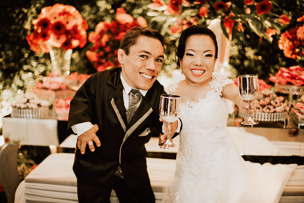 Paulinho e Katyucia em seu casamento, sorrindo para a foto com copos na mão. ele está de terno e ela de vestido branco. há muitas flores atrás deles