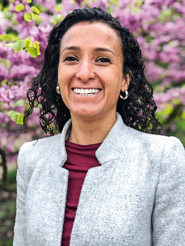 cris foto2.jpg Mulher negra sorri para foto. Ela está em um local com flores roxas atrás dela e cabelos dela voam ao vento