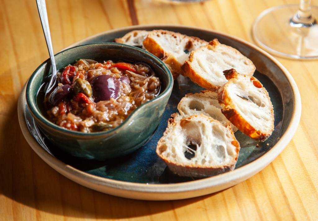 Sobre uma mesa de madeira clara, um prato raso de cerâmica serve de apoio para um recipiente menor de cor esverdeada, também de cerâmica, com caponata (berinjela, pimentão vermelho e azeitonas pretas). Ao lado, algumas fatias de torradas.