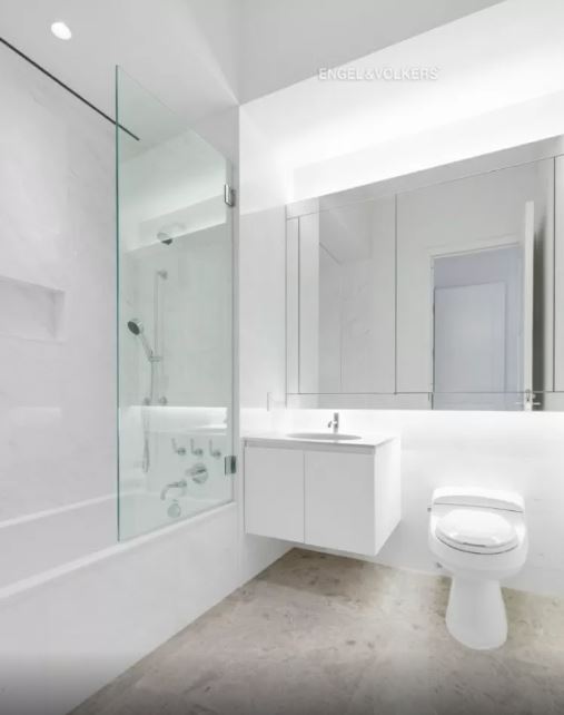 interior-banheiro-432-park-avenue-arranha-céu-milionário-manhattan-problemas-processo-judicial banheiro branco com vaso sanitário à direita, pia ao centro, espelho acima deles e box com vidro e ducha à esquerda