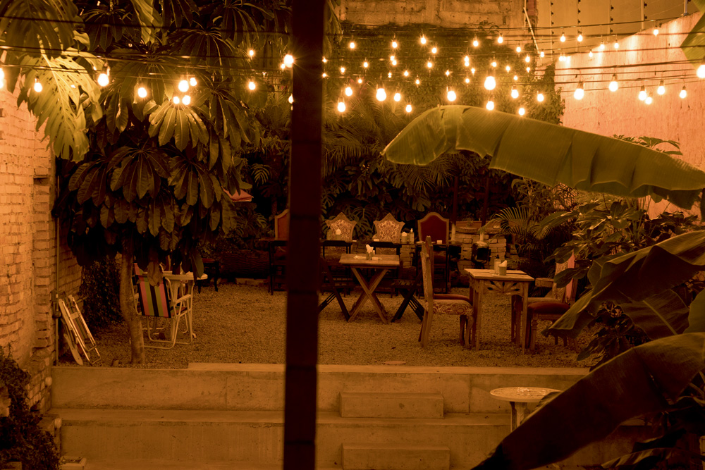 vista noturna, com luzes amarelas dispostas em fios suspensos, do jardim aberto do espaço, com grama, plantas, mesas e cadeiras