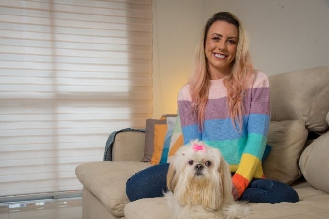 Ligia e a cachorra Maria Cacau sentadas no sofá. lígia está sorrindo para a foto com as mãos na cachorrinha e está usando um sweater arco-íris