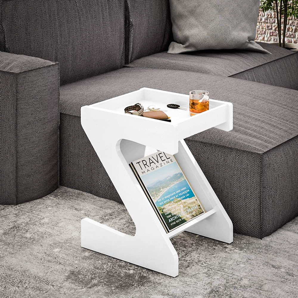 Do lado de um sofá cinza, um móvel de canto de mesa tem uma revista na parte de baixo e uma bebida e acessórios em cima