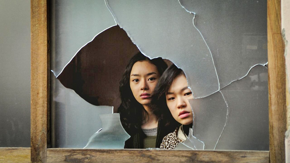 A imagem mostra duas sul-coreanas entre uma janela quebrada, olhando pelo espaço aberto.