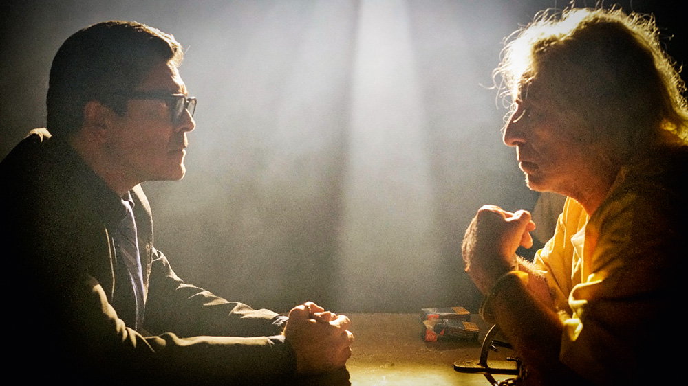 A imagem mostra dois homens, com pouca luminosidade, olhando frente a frente apoiados sobre uma mesa.