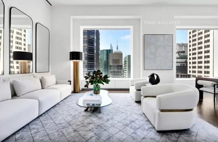 sala-de-estar-2-432-park-avenue-arranha-céu-milionário-manhattan-problemas-processo-judicial sala de estar ampla com diversos móveis claros e duas janelas grandes com vista para a cidade
