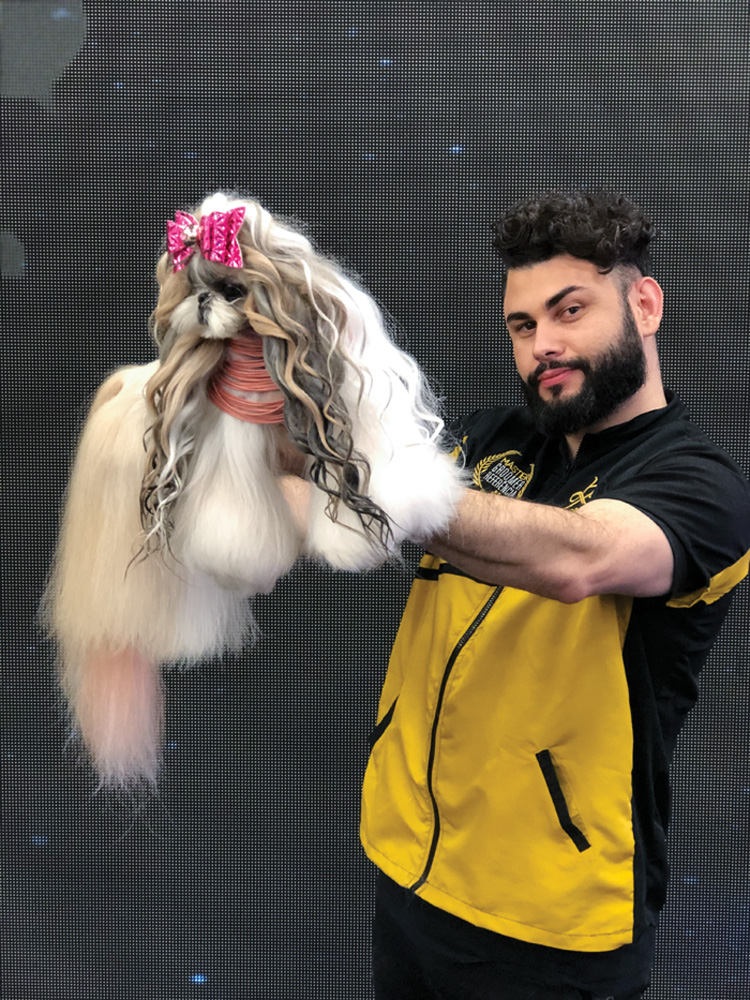 william galharde posando para a foto com carão e segurando no ar com os braços esticados um cachorro que ele acabou de estilizar. o cachorro está com bastante pelo pelo corpo, mas está organizadinho, e está com um laço na testa e colares no pescoço. os pelos que saem da cabeça do cachorro estão trançados e de cores mais escuras que do resto do pelo