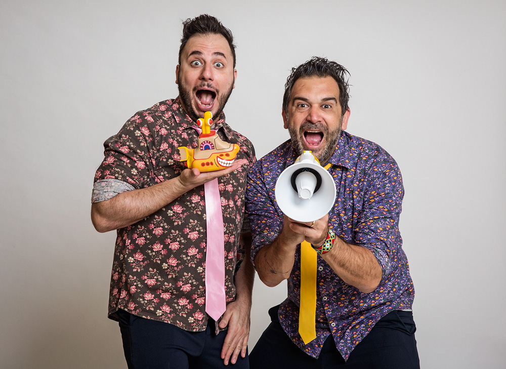 Dois homens vestem camisas floridas e posam para a foto com cara de surpresa. Um segura um submarino amarelo e o outro um megafone