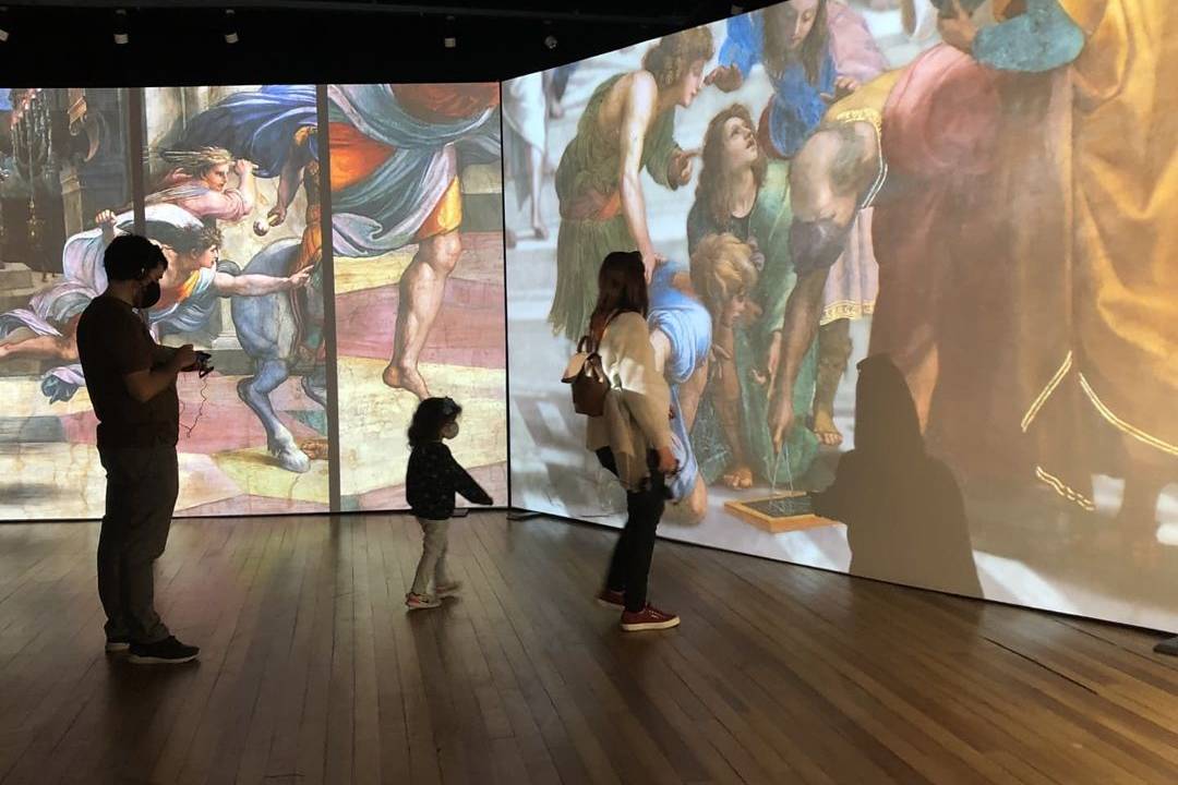 Crianças e mulher em meio à projeção da foto de uma obra de Rafael Sanzio