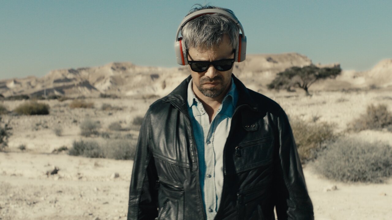 A imagem mostra um homem de fone de ouvido e óculos escuros olhando para baixo em um deserto