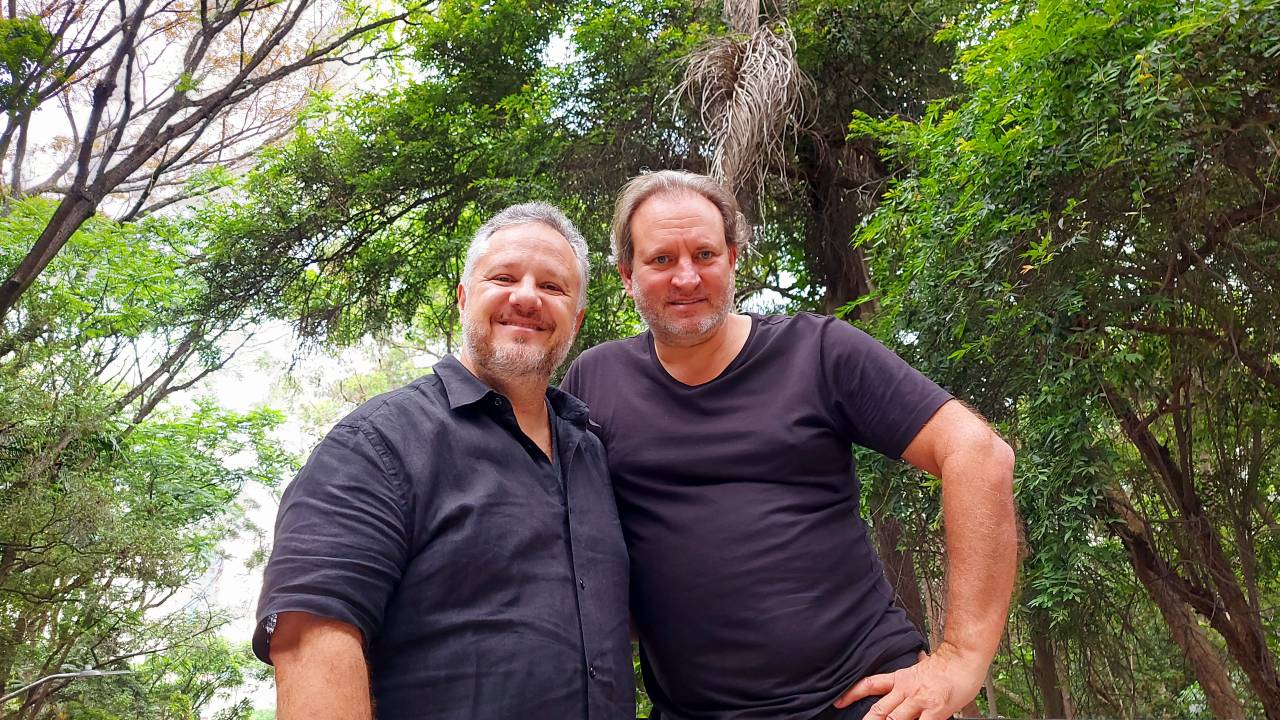 Os empresários Michel Farah e Alexandre Allard posam lado a lado com árvores de fundo, ambos com camisetas pretas.