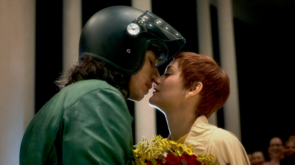 A imagem mostra uma mulher com capacete de motociclista com a parte da frente levantada, quase beijando outra mulher. Ambas estão de olhos fechados, com os narizes se encostando.