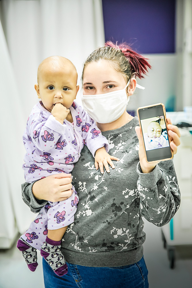A imagem mostra Rita de Cássia segurando sua filha criança Anna Laura. Na mão, um celular mostrando a foto da filha antes do tratamento.