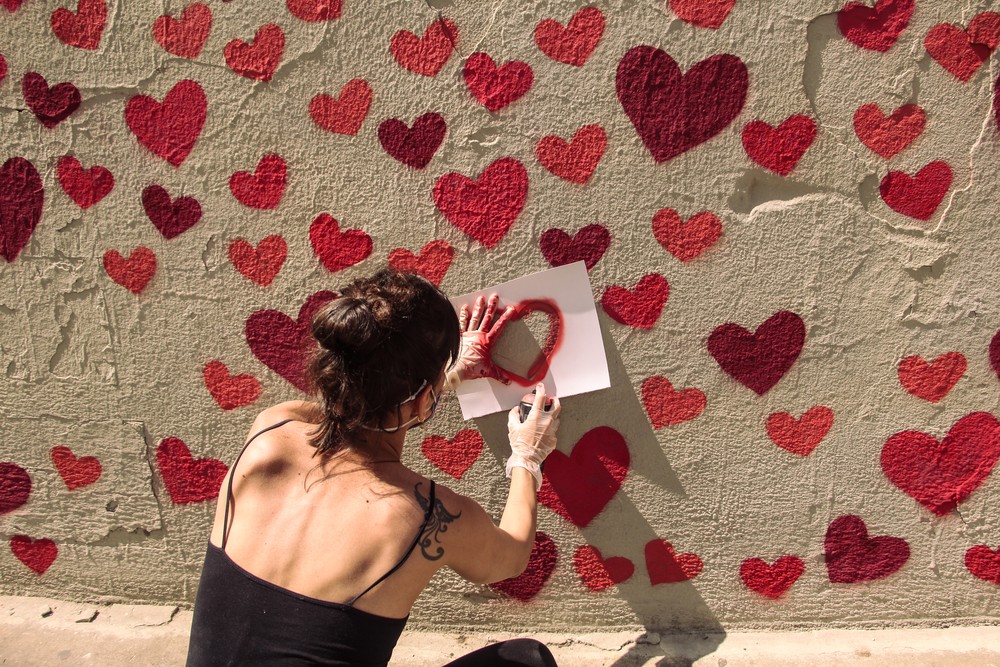 Imagem exibe mulher pintando corações vermelhos com um molde de papel em parede da Praça Roosevelt.