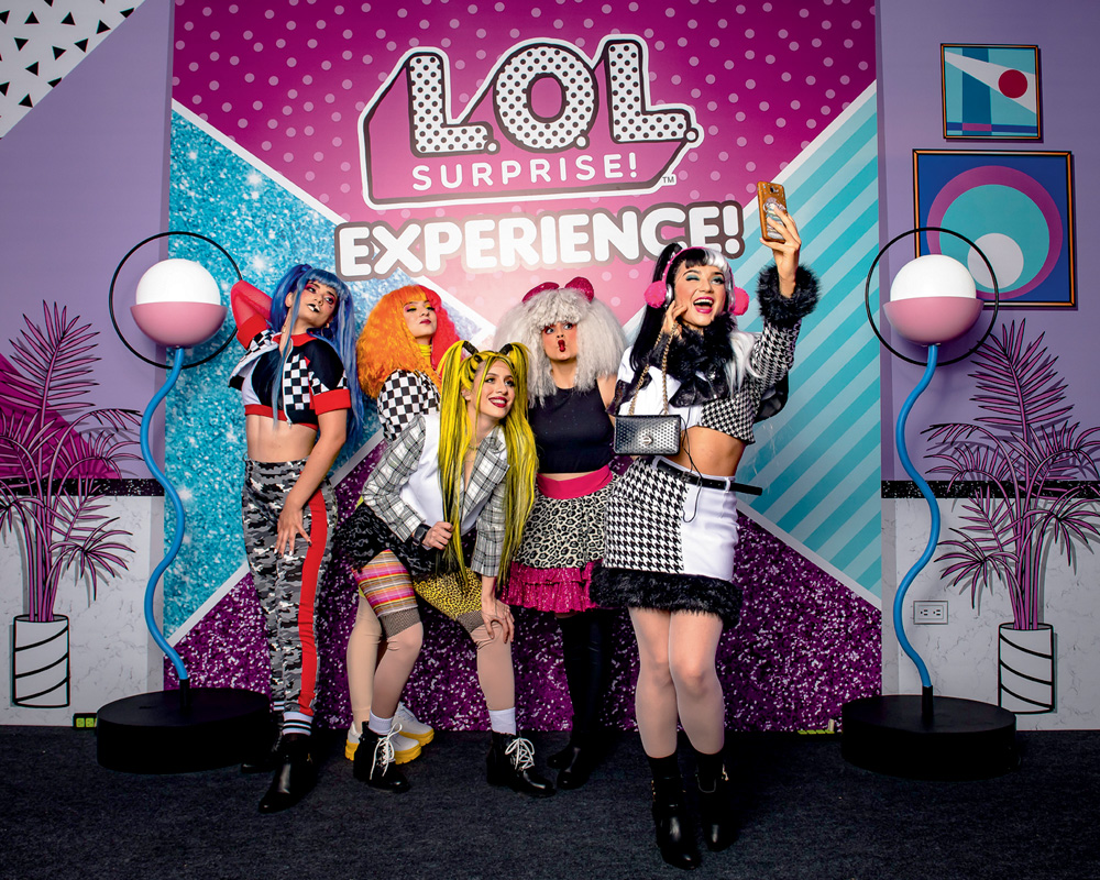 Em um cenário rosa, azul e roxo, cinco mulheres vestidas de bonecas L.O.L. posam para a foto. Elas vestem perucas e roupas chamativas e coloridas. Atrás, um banner escrito 