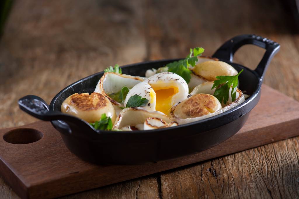 Assadeira de ferro preto com bacalhau, ovo mole e batatas ao murro sobre bancada de madeira.