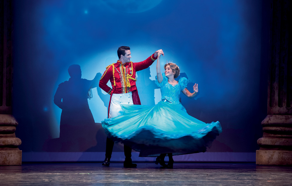 A imagem mostram os dois protagonistas da peça Cinderella. Eles estão dançando, com o príncipe segurando na mão de Cinderella, que está girando em torno de si mesma.