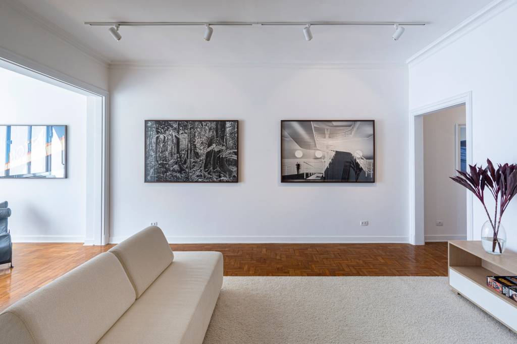 Espaço de galeria de Constance Franciosi, inaugurada em prédio dos anos 1940 no Centro, exibe sala com paredes e sofá brancos. Ao fundo, dois quadros exibem fotografias.