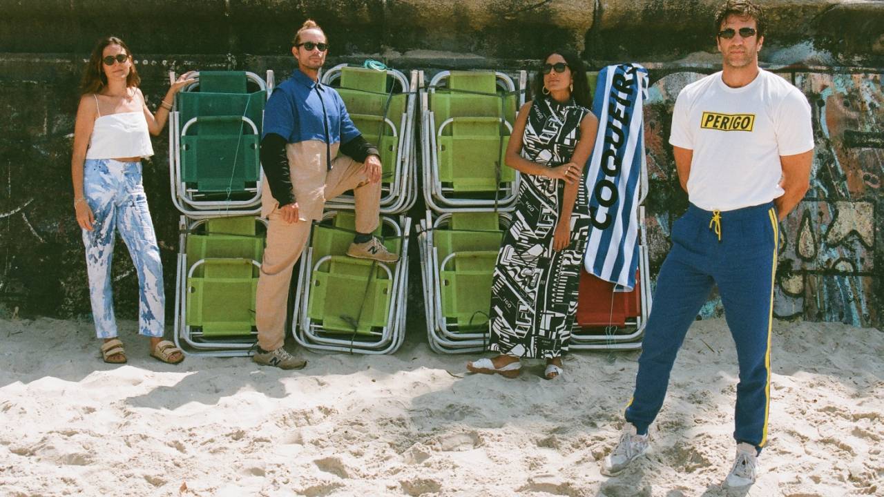 Imagem exibe quatro pessoas vestidas posando na areia, com cadeiras de praia fechadas ao fundo. São dois homens e duas mulheres, todos com óculos escuros e roupas em tons coloridos.