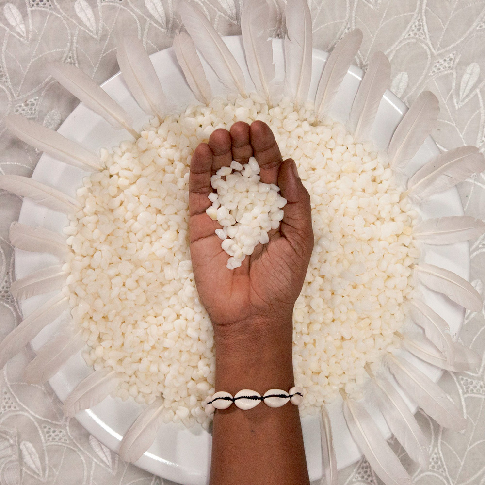 SP Arte A foto mostra um prato branco em uma mesa. Na borda do prato, há penas brancas por toda a circunferência. No interior do prato, há pedrinhas bege/brancas e uma mão negra segura um punhado destas pedrinhas