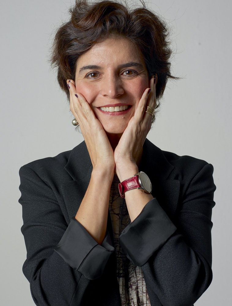 Créditos_BobWolfenson.jpg Mulher posa em um fundo cinza, com roupa social azul escura, cabelos curtos e as duas mãos nas bochechas. Ela está sorrindo
